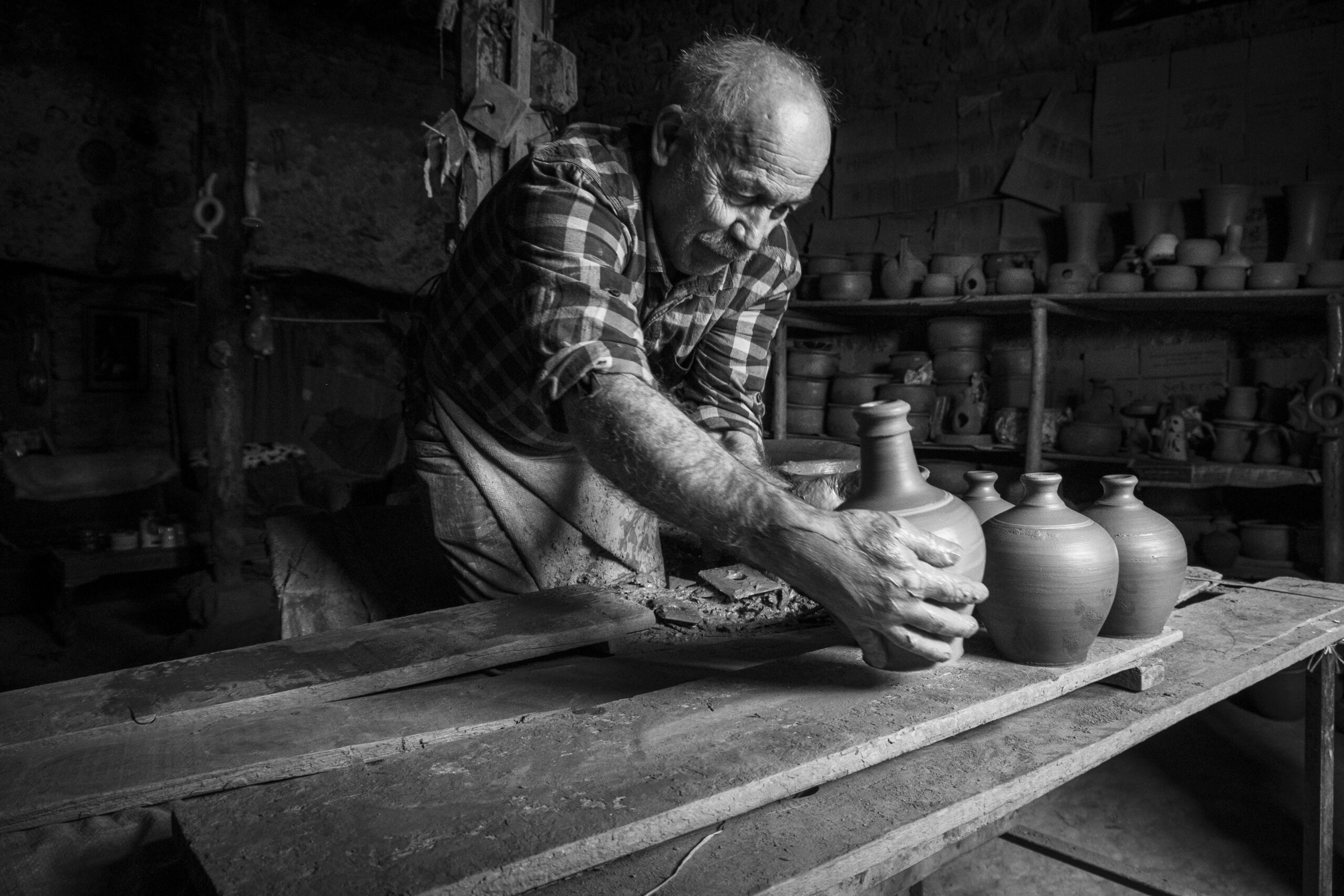 Black and white photo of a senior potter shaping clay pots in a rustic workshop, showcasing traditional craftsmanship.
